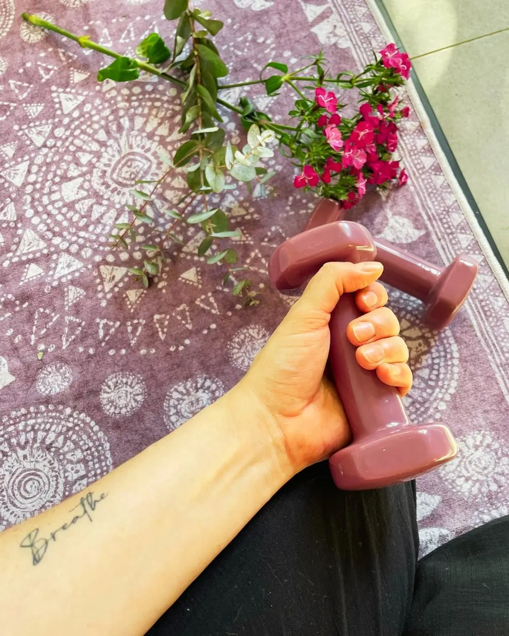 A hand holding a dumbbell beside flowers and a yoga mat